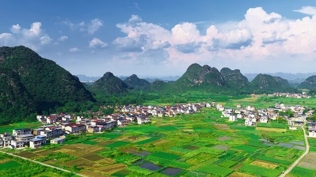 Aerial view of a rural village nestled among green agricultural fields and karst mountain peaks in Guilin, Guangxi under bright sunlight.