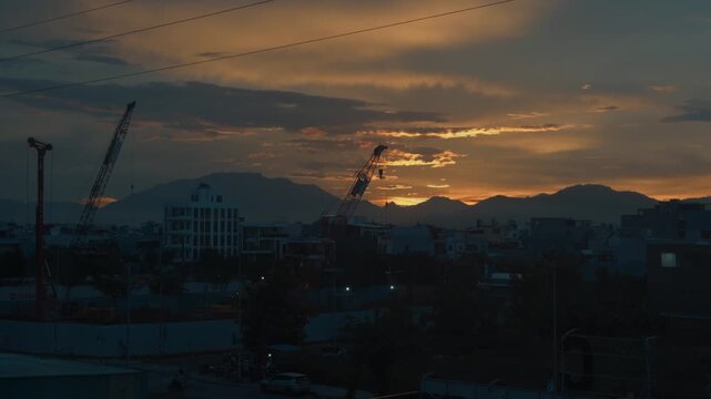 Da nang cityscape views construction cranes silhouetted against a dramatic sunset sky, highlighting urban expansion and modern development with distant mountains creating a serene backdrop at dusk