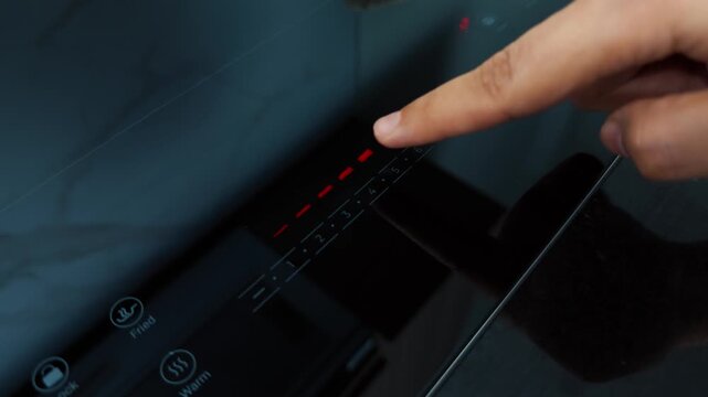 Hand operating a contemporary electric induction cooktop, utilizing the integrated touch panel to precisely control temperature settings for efficient cooking in a modern kitchen environment