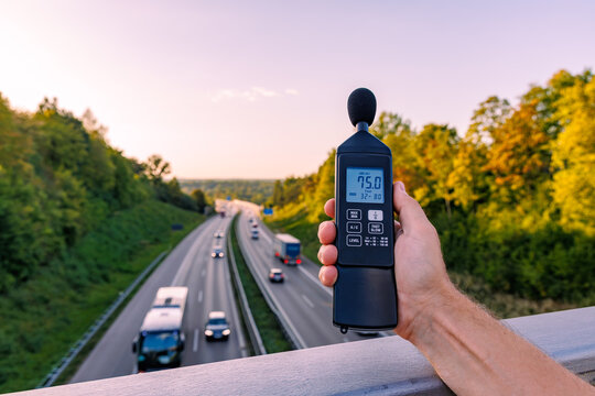 Hand holding digital sound level meter measuring decibel noise over busy highway at sunset, traffic noise pollution and environmental monitoring concept