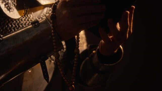 Tilt up shot of young adult knight in medieval armor holding wooden rosary beads and holy book. Clip fits historical documentaries, cinematic trailers, fantasy game teasers, and educational videos