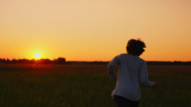 Silhouette of a child running through an open grassy field at sunset, bathed in warm golden light under a tranquil orange sky on a peaceful evening