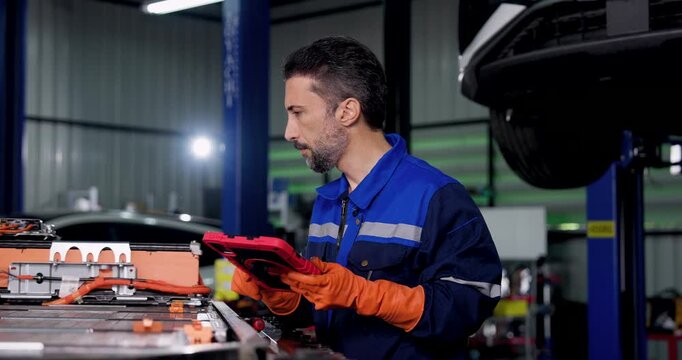 Adult male technician operates digital tablet beside exposed EV battery pack in modern workshop, reviewing vehicle diagnostics during battery inspection and routine electric vehicle maintenance.