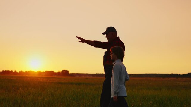 Silhouetted grandfather and child walking together through green field at golden sunset, sharing stories and appreciating peaceful countryside evening landscape