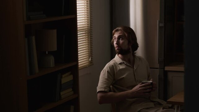 Caucasian young adult man holding mug, sitting and peering through window blinds at home. Clip fits mental health campaigns, loneliness, daily routine storytelling