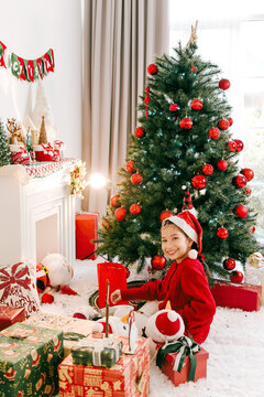 Smiling child in a Santa hat sits by a decorated Christmas tree with red ornaments and wrapped gifts in a cozy living room.