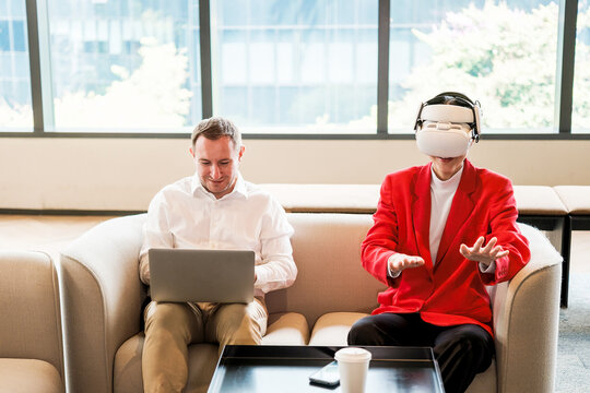 Two colleagues on a couch: one works on a laptop while the other in a red jacket explores virtual reality with a headset, gesturing in a bright modern office lounge.