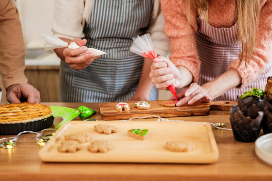 Hands in aprons decorate festive cookies with colorful icing on wooden boards, with a pie and muffins nearby in a cozy kitchen.