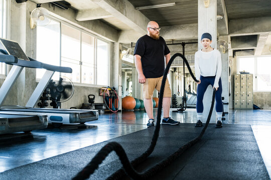 Woman trains with battle ropes under a coach"s guidance in a bright gym with treadmills and fitness equipment