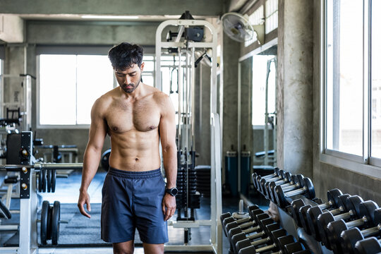 Shirtless adult man stands beside a dumbbell rack in a modern gym, appearing focused after training and conveying strength, discipline, effort and an active fitness lifestyle.