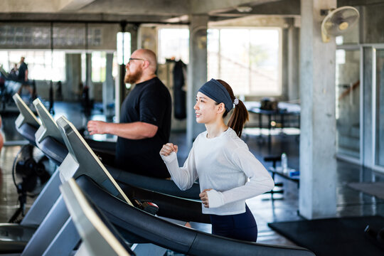 Two people running on treadmills in a modern gym, focused on cardio training amid concrete pillars, mirrors, and daylight.