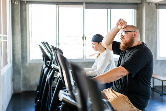 Tired man wipes sweat from his forehead during a cardio workout at the gym while a woman exercises nearby, showing effort, fatigue, endurance and post-workout recovery.