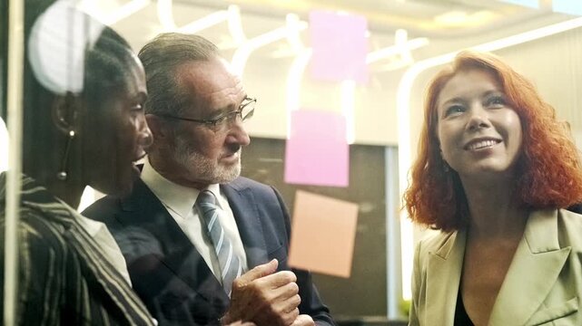 Three business professionals brainstorm with colorful sticky notes on a glass wall in a modern office, sharing ideas and planning strategy in a focused, collaborative team meeting.