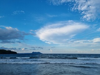 Panoramic view of a serene tropical coastline with rolling ocean waves under a vibrant blue sky featuring scattered soft white clouds at dusk © Fahmi