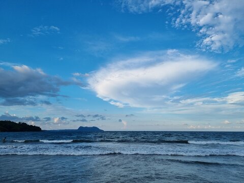 Panoramic view of a serene tropical coastline with rolling ocean waves under a vibrant blue sky featuring scattered soft white clouds at dusk