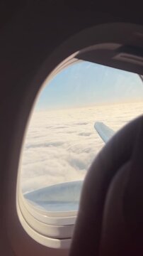 Airplane window view showing wing above fluffy clouds. Blue sky stretches far above white cloud layer. Passenger seat frame partially visible. Image captures peaceful travel moment