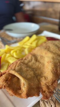 Cachopo, a crispy fried beef and cheese roll, sits on a white plate. Golden fries and a red pepper accompany it. The dish looks juicy and freshly made. Ideal for restaurant menus, food blogs