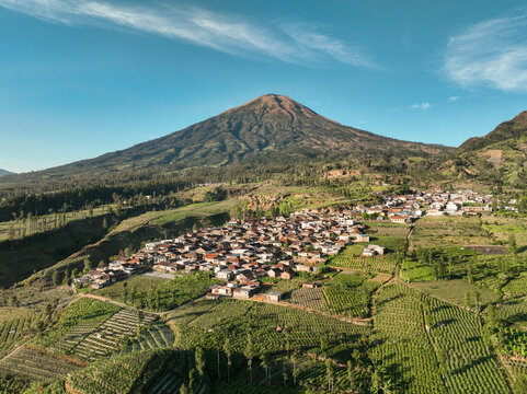 Aerial view of Canggal village nestled among lush agricultural fields and rolling hills at the base of a majestic mountain in Candiroto, Central Java, Indonesia.