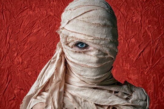 Close-up portrait of person wrapped in textured mummy bandages with one vivid blue eye, dramatic side lighting, and eerie Halloween atmosphere
