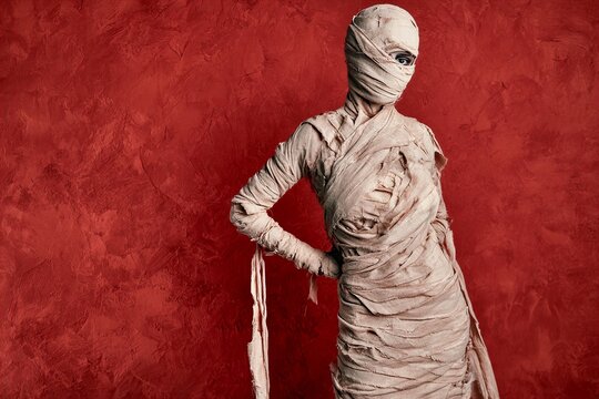 Full body portrait of mummy figure in textured bandages standing against deep red wall with one eye visible