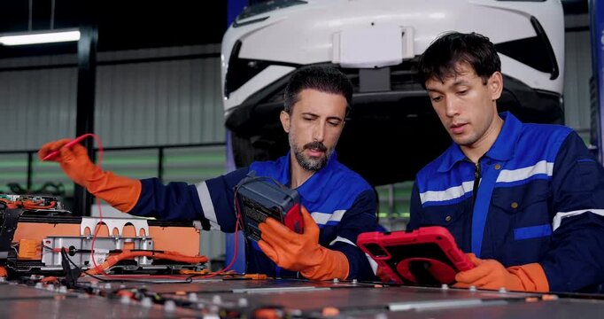 Two adult male EV technicians test exposed high voltage battery pack with meters and probes, comparing readings while coordinating battery diagnostics beneath raised electric vehicle in workshop.