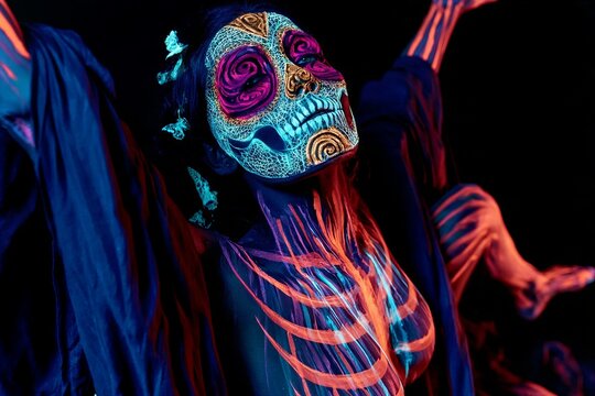 Full body portrait of woman with glowing neon UV skull face paint in ritual pose with raised arms and flowing dark fabric against black background