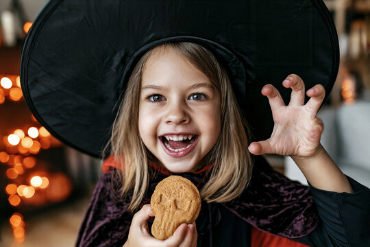 Playful young girl in oversized black witch hat and dark purple velvet cape making claw-like gesture and exaggerated grin while holding skull cookie indoors candlelit