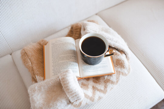 Mug of black coffee on open paper book with folded knitted sweater on couch in living room. Winter cozy atmosphere.