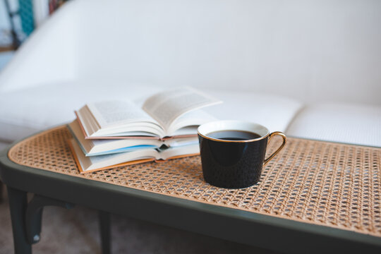 Cup of black tea with stack of open paper books on rattan table in living room close up. Night time.