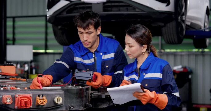 EV technician teamwork with male and female mechanics testing exposed high voltage battery pack, checking connectors and discussing readings during electric vehicle diagnostics in automotive workshop.