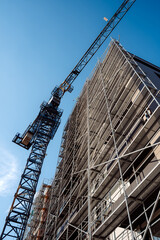 Urban real estate development project showcasing metal scaffolding structures and a heavy-duty tower crane working on a high-rise exterior against a clear blue sky. © Dusko
