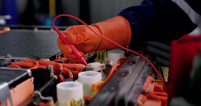 EV battery testing closeup with gloved technician moving meter probe across exposed electrical connection points, performing technical inspection during electric vehicle maintenance in workshop.