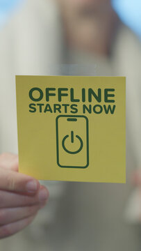 Hand holding a yellow square note with green text offline starts now and a smartphone icon featuring a power button. Symbolizing a digital detox and disconnection from technology for mental well being