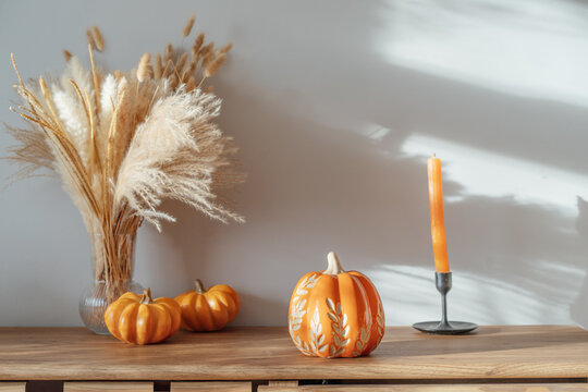 Cozy warm autumn home interior - decorative orange ceramic pumpkins, candles, dried flowers on the wooden console with white wall background. Scandinavian minimalist hygge home fall decor.