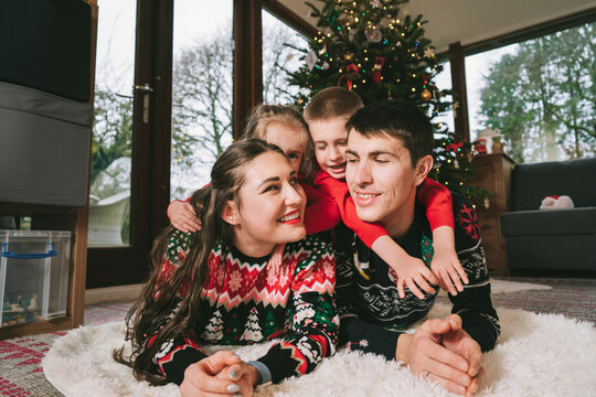 Smiling family portrait in festive sweaters laying under decorated Christmas tree at home during holiday. Happy parents and kids cuddling, feeling excited. Family time together. Happy childhood.
