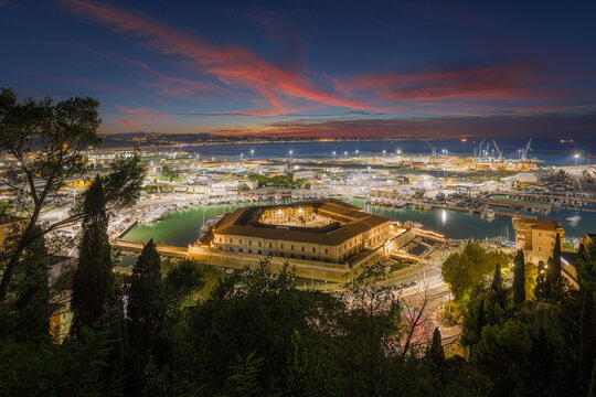 Aerial view of the historic Lazzaretto di Ancona illuminated at sunset within the bustling harbor, featuring industrial port facilities and surrounding greenery in Ancona, Marche, Italy.