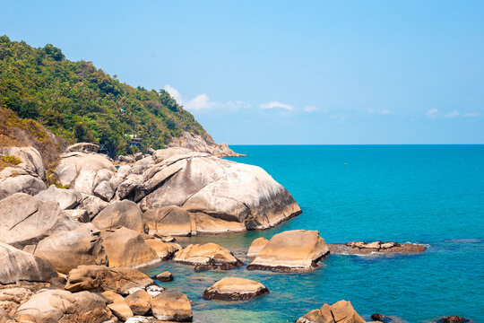 Large stone boulders line a rugged tropical coastline next to bright blue sea water on Koh Phangan. Wild rocky shore landscape under a sunny sky in Thailand. Vacation relaxation, summer getaway.