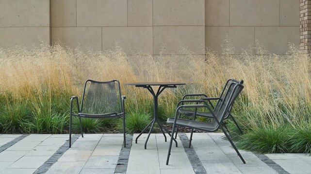 An outdoor seating area features two metal chairs beside a small round table on a stone pathway. Nearby, tall grass adds a natural element to the setting, creating a cozy atmosphere