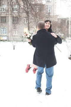 A man lifts a woman in a snowy park while she laughs. They enjoy the cold weather and bright surroundings.