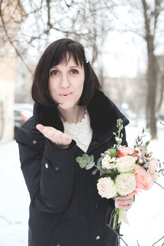 A woman stands in the snow with a bouquet of flowers in her hands. She smiles gently while leaning against a tree.