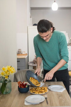 A young man serves food on a table in a kitchen. He holds a pan with cooked items and has plates ready.