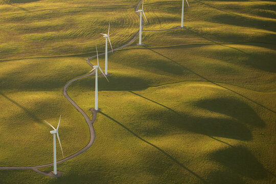 Aerial view of wind turbines standing in rolling green hills with long shadows during golden hour in Rio Vista, California, United States.