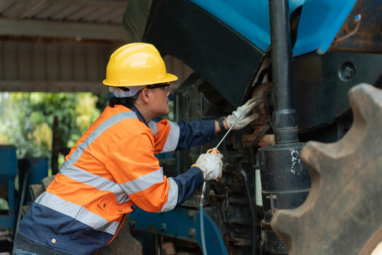 Asian male technician wearing safety helmet using air blower gun to clean heavy tractor engine bay. Professional mechanic maintaining agricultural machinery in garage workshop.