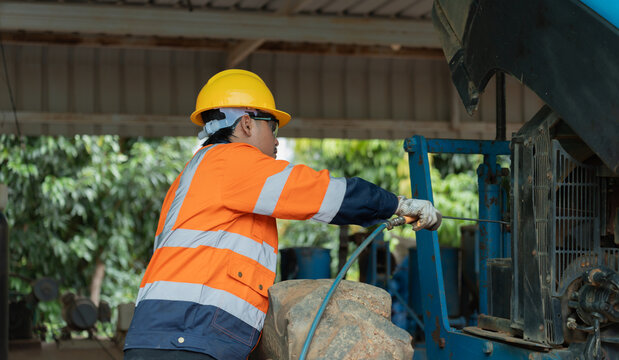 Asian male agricultural technician using air hose gun to blow dust off tractor engine. Experienced mechanic performing routine heavy equipment maintenance in rural workshop.