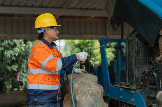 Asian male mechanic in safety helmet using air compressor gun to clean tractor engine. Professional technician maintaining heavy agricultural machinery in garage workshop.