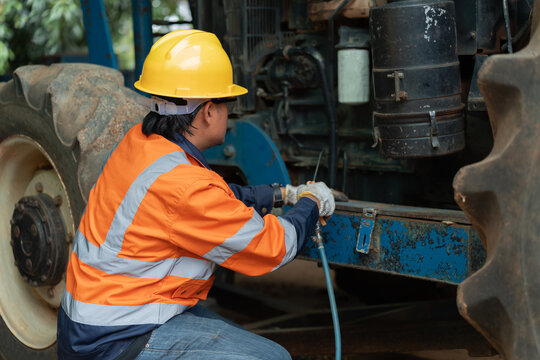 Asian male engineer squatting near big tire to clean tractor engine with air blower gun. Professional worker carrying out maintenance on heavy farm machinery outdoors.
