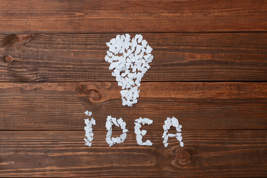 Conceptual image. lightbulb shape and the word IDEA, meticulously crafted from small, torn white paper pieces. Set against a rich, dark brown wooden background with visible grain. Overhead view.