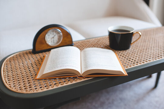 Open paper book with cup of coffee and table clock in living room close up. Reading hobby.