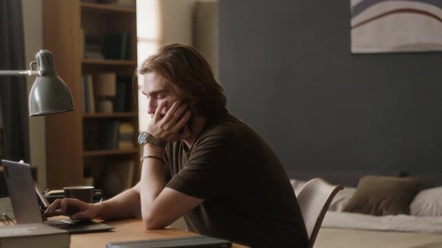 Young adult Caucasian man drinking coffee and working on laptop with tired expression at desk. Clip suitable for remote work burnout, freelance fatigue and corporate mental health campaigns