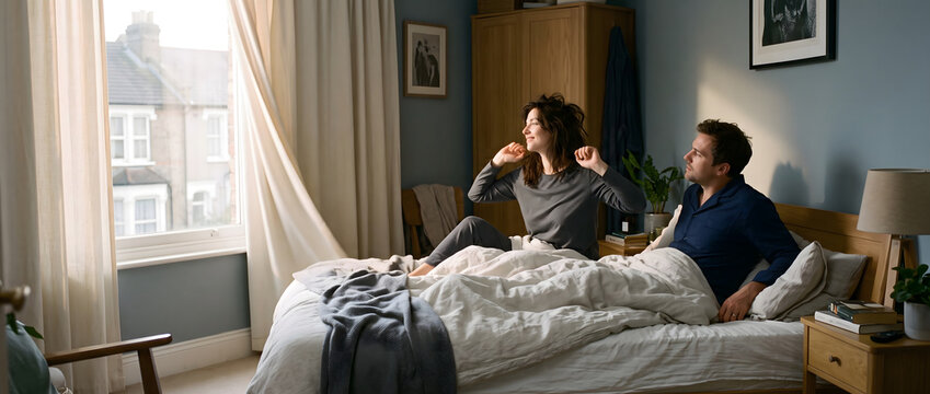 A man and a woman wake up in a sunlit bedroom, sitting on a bed covered with bedding, in casual sleepwear.